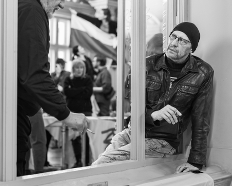 Czarno-białe zdjęcie. Aktorzy Tomasz Międzik i Tomasz Wysocki znajdują się przy oknie w ozdobnym wnętrzu teatru. Palą papierosy, jeden pochyla się lekko do przodu, drugi siedzi na parapecie, oparty o ramę okna. W tle widać inne osoby oraz fragment flagi.